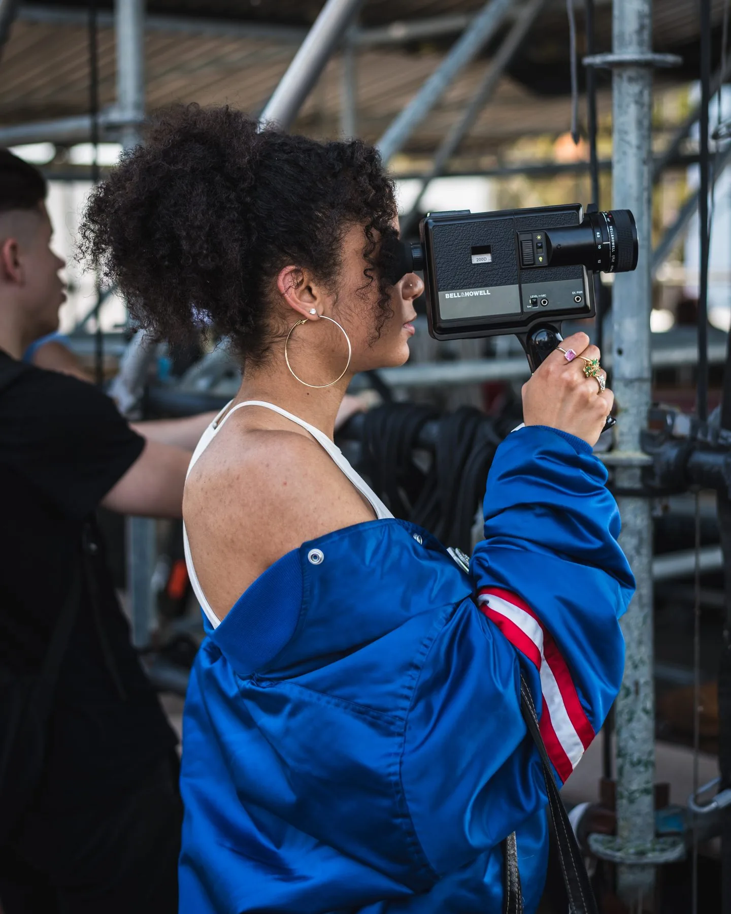 A crew member filming on a Bell and Howell Super 8 camera against stage scaffolding.